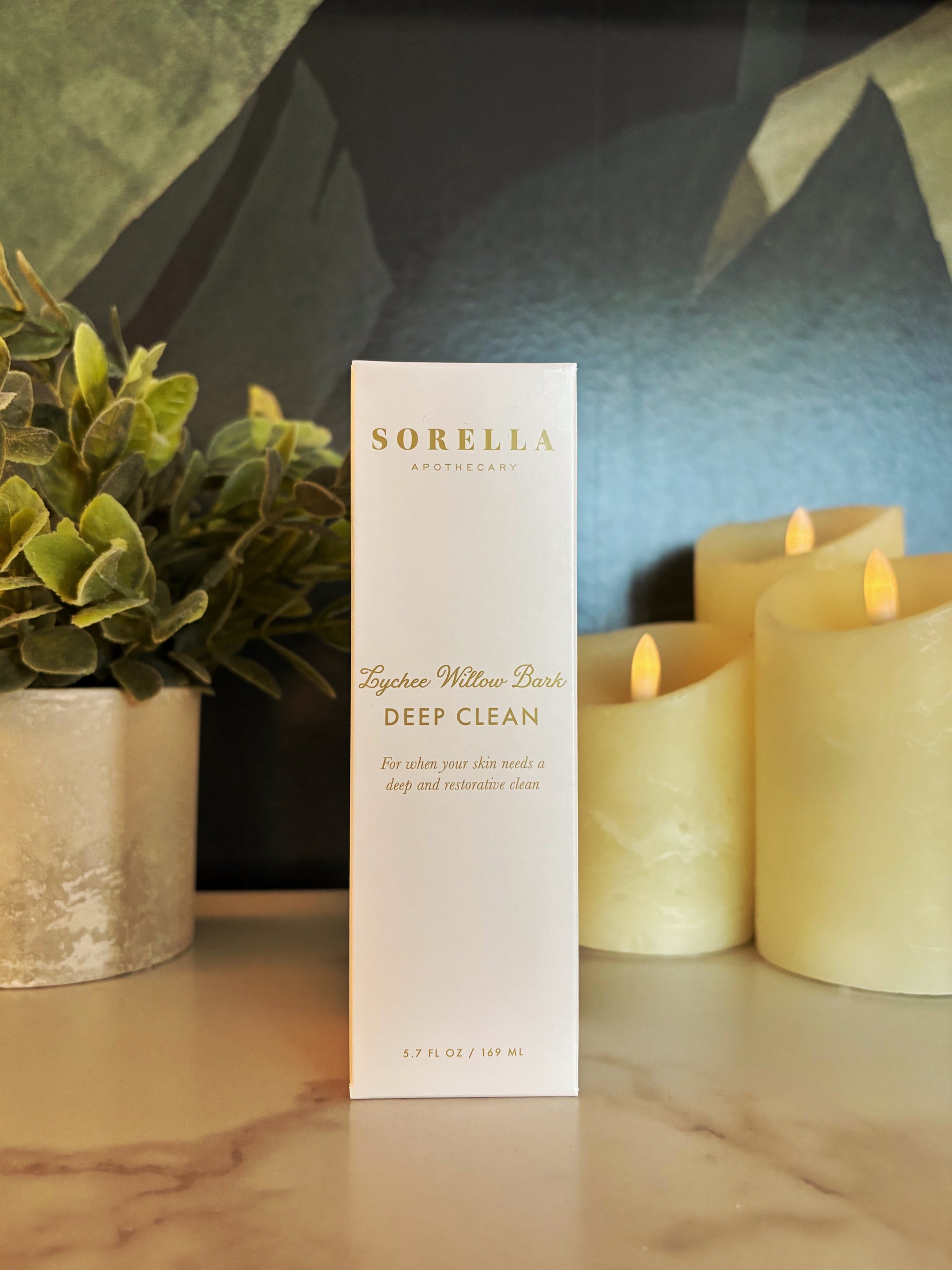 A tall white box for Sorella Apothecary 'Deep Clean' lychee willow bark cleaner stands on a marble surface, styled with a potted plant on the left and lit candles on the right.