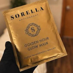 A hand wearing a black glove holds a gold foil packet of Sorella Apothecary Golden Hour Glow Mask against a softly blurred indoor background.
