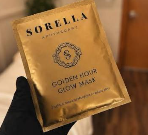 A hand wearing a black glove holds a gold foil packet of Sorella Apothecary Golden Hour Glow Mask against a softly blurred indoor background.